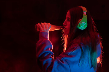 Beautiful young woman drinking a beer with headphones in dark background with neon color lights.Night Club party lifestyle with copy space.