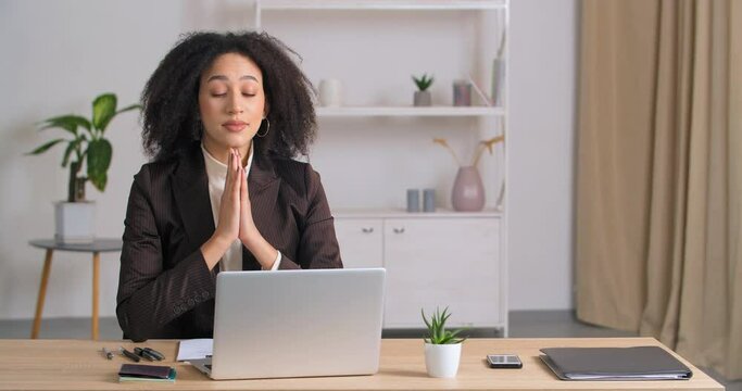 Afro american curly-haired business woman sitting in office folding her palms in supplication asks god for luck in new project, young ethnic student making request gesture hopes for good test results