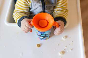 Young toddler child learning to drink out of a training cup; seated in high chair wearing yellow...