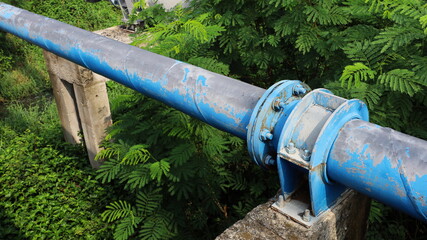Clamps and nuts to hold old pipes on posts. A blue main water supply pipe with clamps and bolts connecting pipes on a concrete column base across the water canal with green plants. Selective focus