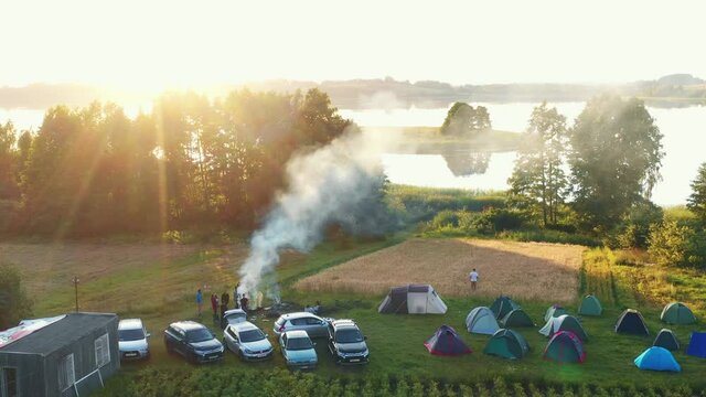 Drone flying above camping spot with fire, cars and tents towards large lake and epic summer sunrise lens flare panorama