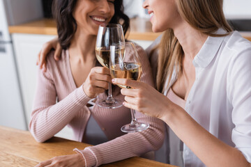 cropped view of happy lesbian couple clinking glasses of white wine in kitchen