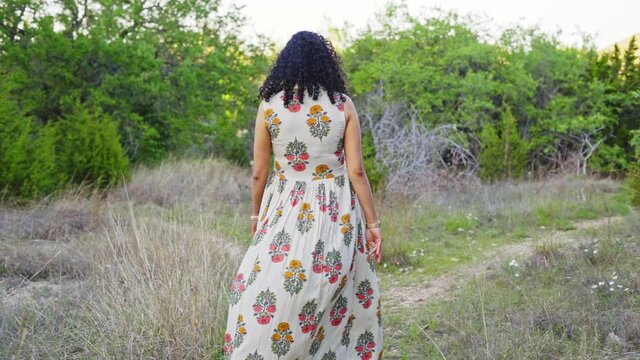 Woman with curly hair in a flowy sun-dress walks on a path that ends in a fork to the left and right.