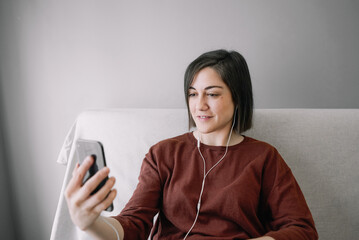 Woman having a video call with her black cell phone and white headphones and sitting on the sofa near the bright window in her living room at home 