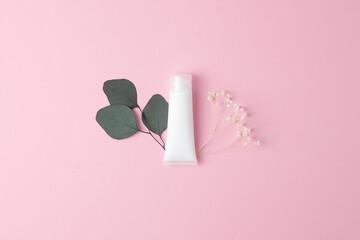 White tube of cosmetic cream with flowers and green leaves on pink background. Flat lay, copy space