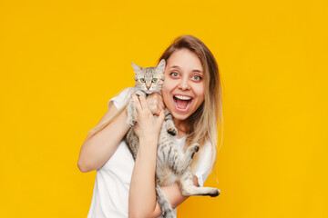 A young pretty caucasian excited smiling blonde woman in a white t-shirt with a cat in her hands is happy about the news isolated on a bright color yellow background. Excited teen girl with kitten