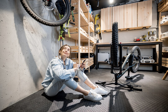 Young relaxed handywoman sitting with phone while repairing her bicycle in the well equipped workshop at home. DIY concept