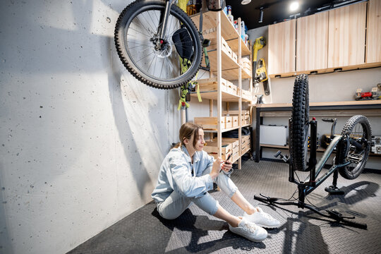 Young relaxed handywoman sitting with phone while repairing her bicycle in the well equipped workshop at home. DIY concept - Powered by Adobe