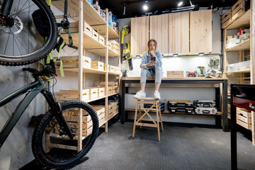 Well equipped home workshop with bicycles, wooden shelves and woman sitting on a workbench - obrazy, fototapety, plakaty