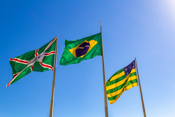 Três bandeiras hasteadas e céu azul ao fundo. Bandeira do Brasil, do Estado de Goiás e da cidade de Goiânia.