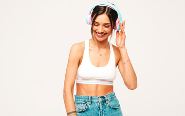 Beautiful smiling woman dressed in jeans clothes. Sexy carefree model listening music in wireless headphones. Adorable and positive female posing on grey background in studio