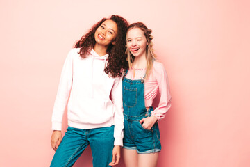 Two young beautiful smiling international hipster female in trendy summer clothes. Sexy carefree women posing near pink wall in studio. Positive models having fun. Concept of friendship