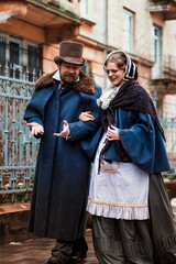 Couple wearing traditional victorian clothes, man and woman walking and talking. Fashion and trends of previous epochs and times. Male wearing cloak, female in dress and hat.  - obrazy, fototapety, plakaty