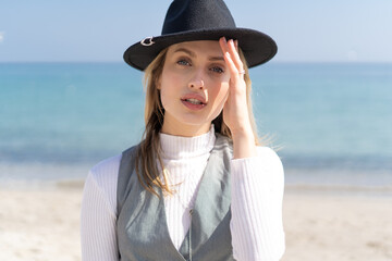 portrait of a stunning young woman holding her fashion hat and looking in the camera with a beautiful blue eyes and blonde hair on a beach 