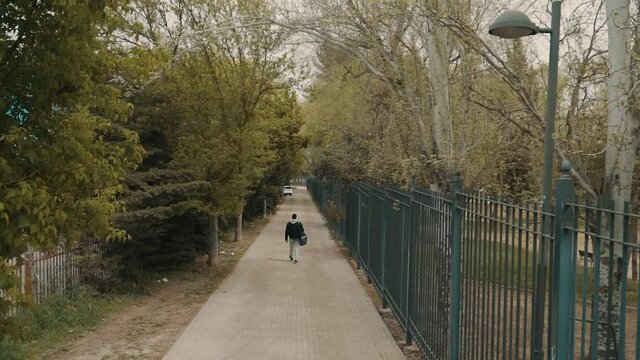 Vista a&eacute;rea de un  hombre caminando por el parque, hombre caminando con su bolsa de deporte, hombre en ch&aacute;ndal gris  y chaqueta negra caminando por la acera , vista superior desde los arboles