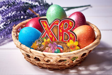 A small basket with colorful painted eggs and a bouquet of lavender on a white wooden background. Russian letters 