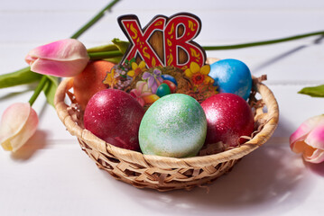 A small basket with colorful painted eggs and a bouquet of tulips on a white wooden background. Russian letters 