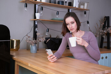 Beautiful young brunette smiling woman in casual clothes with cup of tea using mobile phone