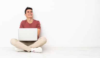 Teenager man sitting on the flor with his laptop looking up while smiling