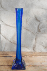 A tall, thin blue glass vase for flowers stands on a wooden base against a plastered wall.