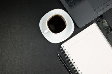 Desk workspace with blank notepad, pen, glasses, laptop and coffee cup on black background. Top view with copy space