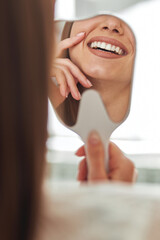 Happy young woman smiling checking out her perfect healthy teeth in the mirror close up, at the dentist office
