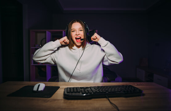 Joyful beautiful female gamer won an online game, rejoices with a raised hand and smiles with joy. Streamer winner plays online games at home on the computer