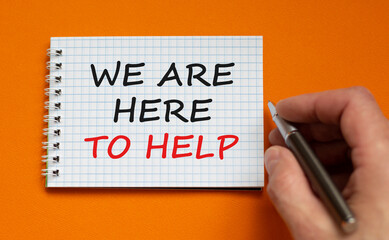 Support symbol. White note with words 'we are here to help'. Businessman hand with pen. Beautiful orange background, copy space. Business and support concept.