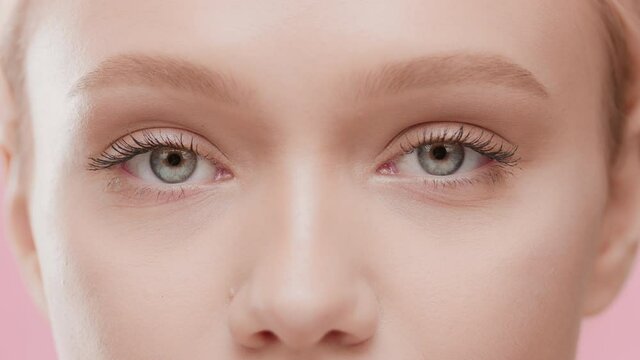 Extreme close-up of attractive Caucasian female model with golden hair opens her eyes moving her head up and looking at the camera against pink background | Face care commercial