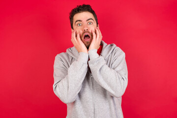 Stupefied Young caucasian man wearing tracksuit over red background expresses excitement and thrill, keeps jaw dropped, hands on cheeks, has eyes popped out