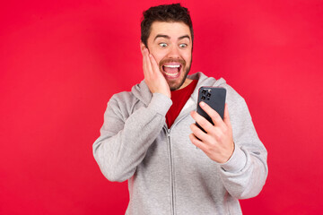 Young caucasian man wearing tracksuit over red background holding in hands cell reading sms using new app 5g