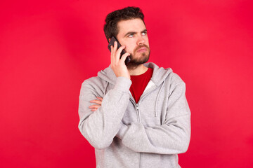 Sad Young caucasian man wearing tracksuit over red background talking on smartphone. Communication concept.