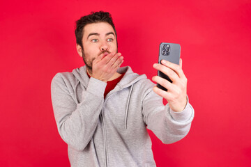 Young caucasian man wearing tracksuit over red background blows air kiss at camera of smartphone and takes selfie, sends mwah via online call.