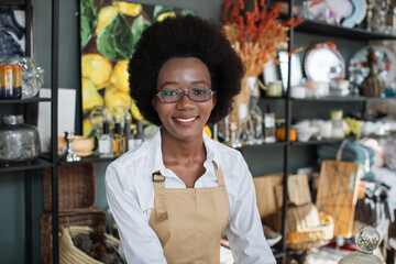 Close up of smiling confident African female worker in beige apron, posing at camera during work at decor shop. Stylish home decorations on the shelves