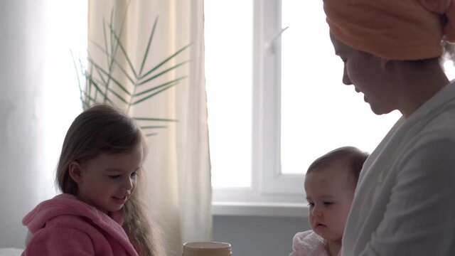 Authentic cute young woman applying face mask of baby girl. mom with 2 children daughters different ages in bathrobes after bath on white bed oposite window. Spa, motherhood, childhood, family concept
