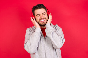 Pleased Young caucasian man wearing tracksuit over red background enjoys listening pleasant melody keeps hands on stereo headphones closes eyes. Spending free time with music