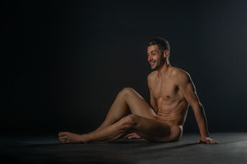 Fashion nude photo of a male model with seductive figure sitting isolated on the floor in a studio