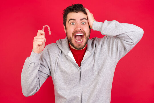 young handsome surprised caucasian man in sports clothes against red wall holding an invisible aligner braces and holding hand on his head. Big sales and promotion.