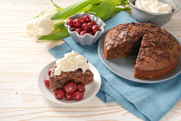 Homemade chocolate cake and bowls with morello cherries and whipped cream, some tulip flowers and a blue napkin on a bright wooden table, copy space
