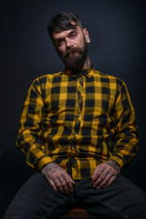 Obraz premium Tattooed man with a beard wearing checkered shirt is posing seated on a chair in a studio
