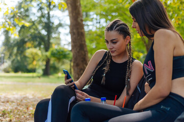 Beautiful young female friends sitting on a bench in a park while looking at a smartphone.