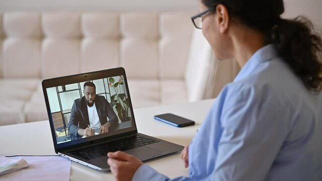 Online business meeting via video conference. View over shoulder of a business woman to a laptop screen with African American colleague, coworkers talking using an online communication application