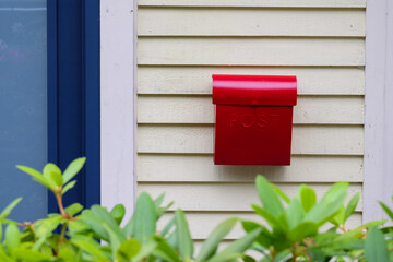 The exterior of a yellow clapboard wall of a house with a bright red metal mailbox. The postal container is to the right of a blue door with glass.  There are some green shrubs in the foreground. 