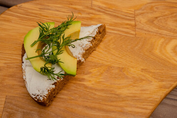 A sandwich of black bread with avocado, cheese and dill is placed on a wooden board