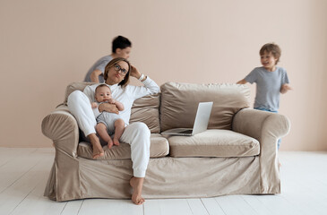 Young single mother working on laptop in loft sitting on couch while her sons running around her and shouting. High quality photo