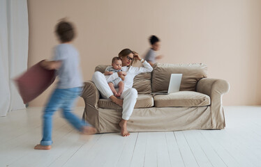 Young single mother working on laptop in loft sitting on couch while her sons running around her and shouting. High quality photo