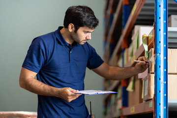 Male employee warehouse worker using clipboard checking products or parcel goods on shelf pallet in...