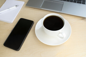 A white cup of coffee and a black mobile phone sat next to a gray laptop on a wooden table. Online Business and Education concept