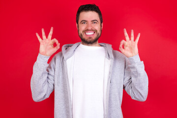 Glad young handsome caucasian man in sports clothes against red wall shows ok sign with both hands as expresses approval, has cheerful expression, being optimistic.