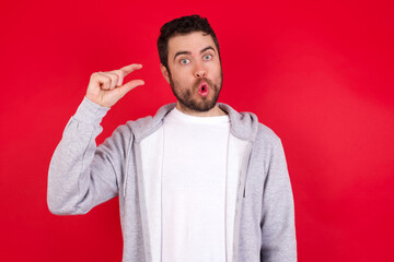 Shocked young handsome caucasian man in sports clothes against red wall shows something little with hands, demonstrates size, opens mouth from surprise. Measurement concept.